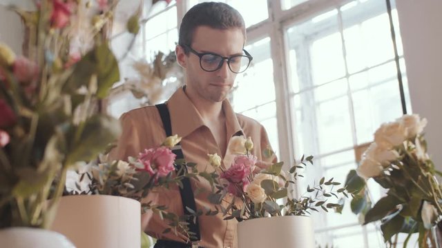 Low angle view of young handsome male florist in glasses cutting rose with shears and putting it into hatbox while making beautiful floral composition in flower shop