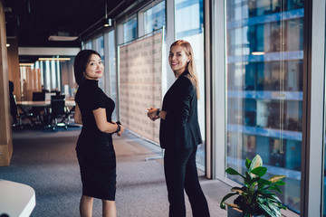 Young women employees in formal wear standing in office satisfied with friendship on work talking...