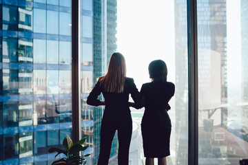 Back view of female colleagues in formal wear standing near window looking at modern exterior of...