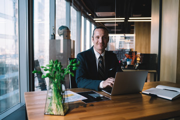 Cheerful prosperous male entrepreneur in formal wear keyboarding on laptop computer satisfied with...