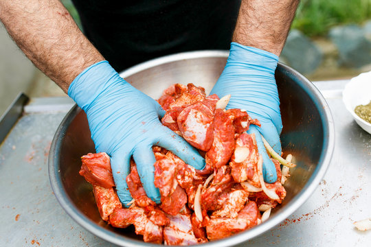 Close-up. Male hands in disposable blue gloves mix red meat in spices in a metal plate. Red meat is mixed in a barbecue marinade.