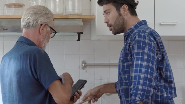 4K Caucasian family handsome adult son helping his senior father solving cooking problem from online recipe in smartphone in the kitchen. Dad and son spending time together at home