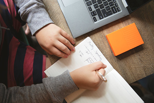 A teenage boy is calculating his budget per month using a notebook and laptop at his desk