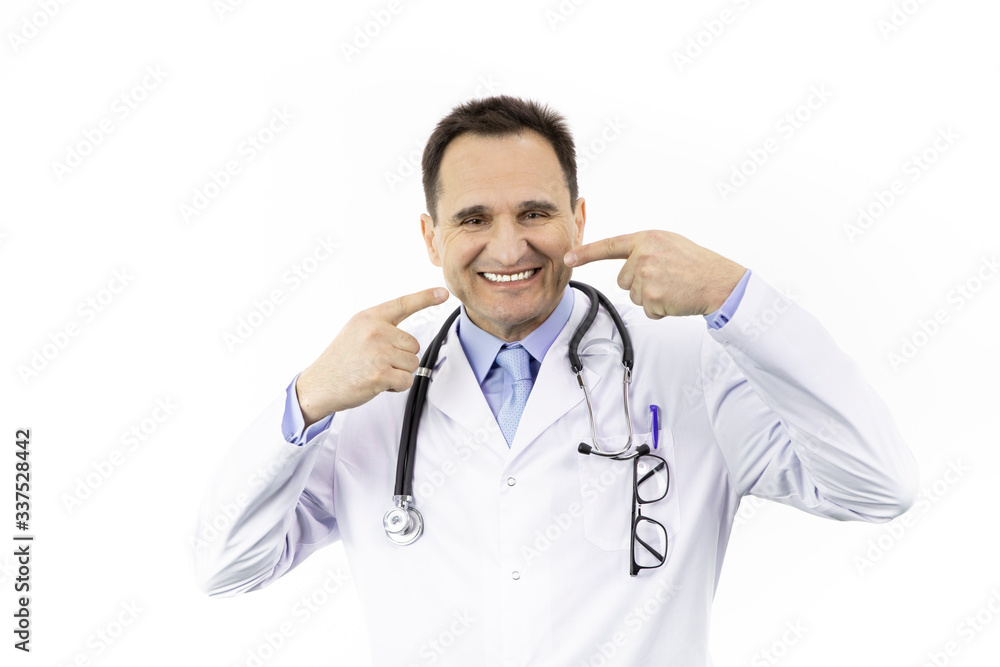 Wall mural Close up portrait of happy with snow-white smile adult male doctor pointing on her perfect clear white teeth, isolated on white background. Physician in medical gown with stethoscope over neck.