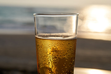 Glass of a cold beer at sunset, sea and beach in the background.