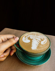 There is a cup of cappuccino on Kraft paper and it is held by a man's hand, at the top of the frame there is plenty of space