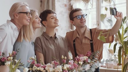 Young male florist in apron smiling and posing for smartphone camera while taking group selfie with cheerful female colleagues during workday in flower shop