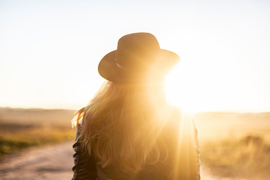 Girl in sunny light with long hair in black hat on beautiful sandy road looks at sunset. Outside. View from backside. Warm sunny day. Travel concept. New life, new horizons and opportunities