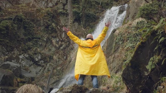 Young male traveler in a yellow raincoat enjoys a beautiful waterfall. Hiking in the mountains. The hiker runs to the waterfall, raises his hands up. Enjoys lifestyle. Concept: religion, bio, ecology