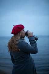 Beautiful pregnant caucasian girl with large belly and long curly hair in coat and red beret drinking coffee from thermocup blue sea nature backgroud. Maternity lifestyle shoot. Life after baby birth
