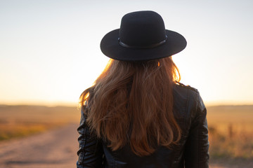 Girl with long brown hair in leather jacket and black hat on beautiful sandy road looking at sunrise. Outside. View from the backside. Warm sunny day. Travel concept