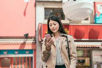 Happy woman smiling and standing in street using smartphone. young girl in smart casual wear with cellphone looking online map in dotonbori. lady finding famous restaurant online on mobile phone