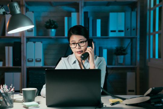 business deadline and technology concept. asian japanese businesswoman with laptop computer calling on phone at night office. lady worker having discussion with colleagues during overtime working.