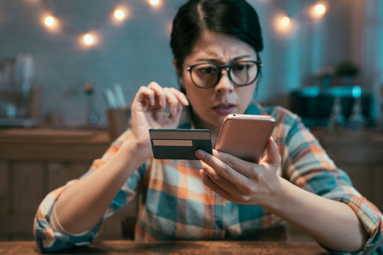 Confused asian chinese woman paying online with credit card sitting in dark home kitchen at night. shocked frowning female holding debit card and cellphone having no money or blocked bank account.