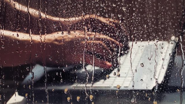 Close up. The guy is working alone at a laptop, in the foreground a window with drops after the rain. Typed on the notebook keyboard Illuminated sunlight. Remote work from home during the quarantine