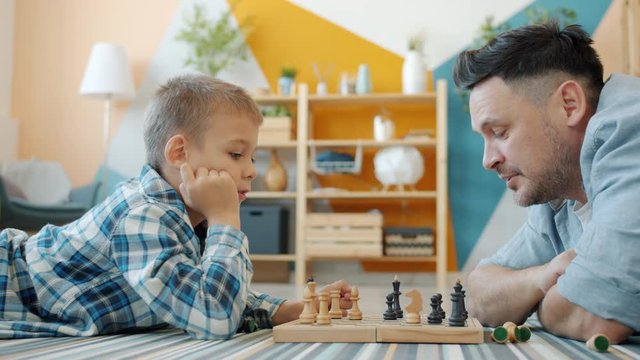 Little kid enjoying chess game with caring father spending leisure time at home playing together on floor. Family activities and fatherhood concept.