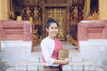 Fotobehang Portrait of Asian woman visiting Wat Sensoukaram in Luang Prabang, Laos. Portrait of woman wearing laotian costume and holding wooden bamboo contained sticky rice inside. © boyloso