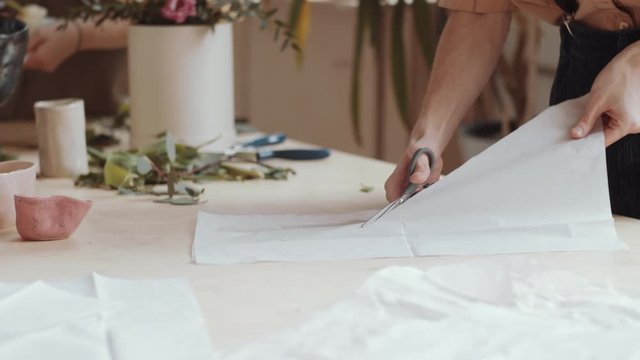 Tilt up shot of young professional male florist in apron and eyeglasses cutting wrapping paper with scissors while working at table in flower shop