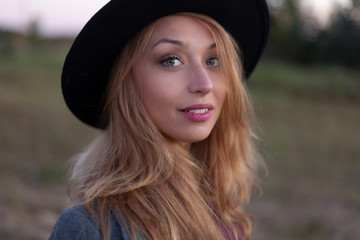 Close up portrait of beautiful caucasian woman with long fair hair in black hat looking at camera outside. Green nature backsides. Back to basics, relaxing concept