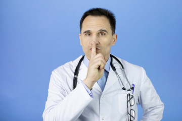 Portrait of a Caucasian adult doctor in a white coat with a stethoscope over his neck on a blue background. Index finger at the mouth. Keep quiet. Close-up.