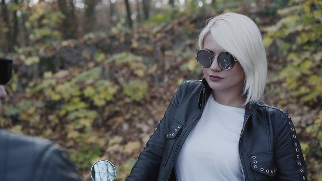Portrait of proud blonde in sunglasses stands at motorbike in park