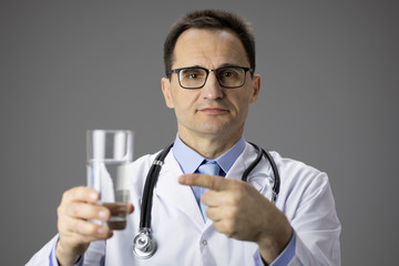 A physician in a white coat with a stethoscope on his neck holds a glass of water in his hands and points a finger at it. Healthy lifestyle. Thirst. Proper nutrition. Water-salt balance. Close-up.