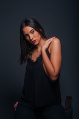 Fototapeta na wymiar Sexy brunette wearing a black tank top and black pants posing while sitting on the top edge of a chair inside a studio on a dark background