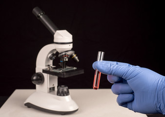 Microscope blurred and hand with surgical glove holding vial with red solution. Black background with space for text
