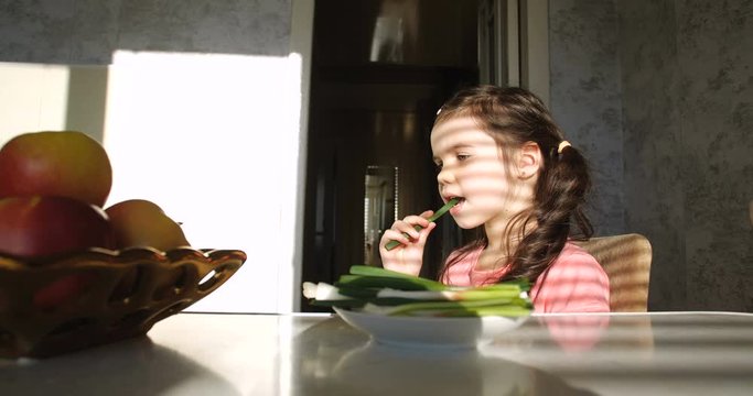 Little girl of five years old sitting in the kitchen at table, taking bitter and healthy green onions from plate and eating with appetite. Concept of unusual eating habits and healthy vegetarian diet