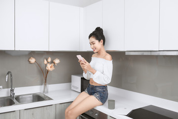 Asian woman At Home Sitting On Kitchen Island Whilst Using Mobile Phone and drinking coffee