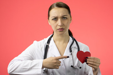 Beautiful serious female cardiologist in white medical coat and stethoscope pointing on red paper...