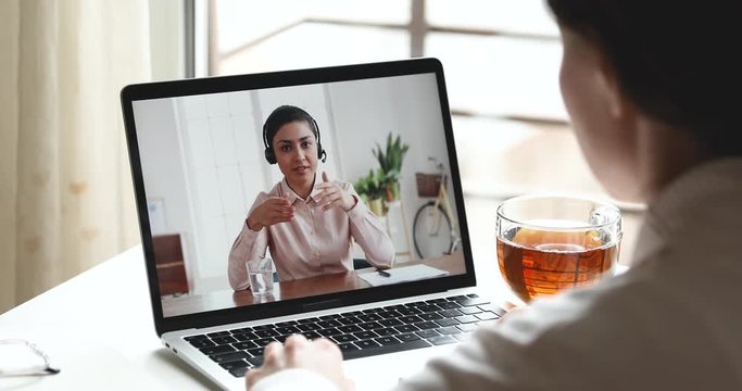 Indian female online teacher, customer service support manager wearing headset consulting client, student, remote worker in webcam video call virtual conference on laptop screen. Over shoulder view