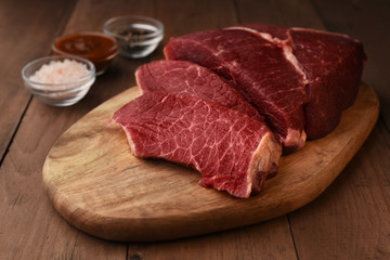 Angus beef meat sliced on a wood cutting board. Raw red meat on a board and seasonings around on wooden table. Salt, peppercorns and spicy red tomatoes sauce. Ready to cook at home. 