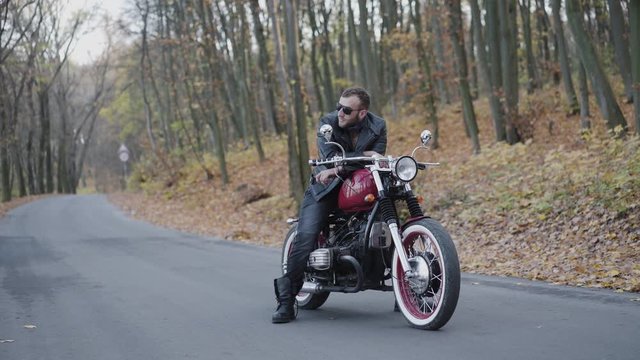 Portrait of brutal biker behind a motorcycle stands among park roadway