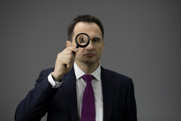 Concentrated male adult businessman in formal suit looking through loupe, isolated grey background. Copy space. Portrait of handsome man analysing information, statistics and business statement