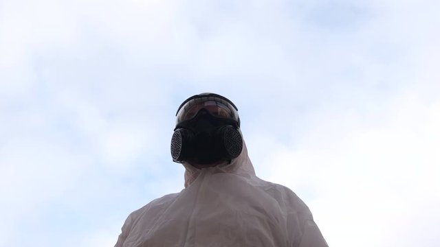 Man wearing protective suit, gas mask and goggles as protection against virus infection, outside from below against blue sky. Turns slowly.