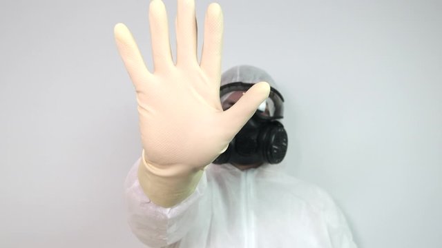 Male wearing protective suit, gas mask and goggles as protection against virus infection raises hand in stop gesture. Slow motion.