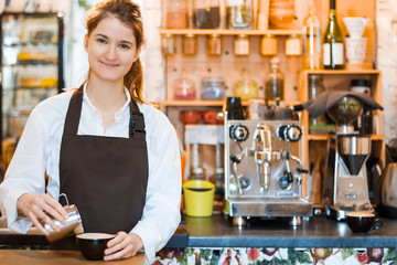 Woman is pouring hot milk and froth from metal jug to cup. Process of preparing, brewing americano,...