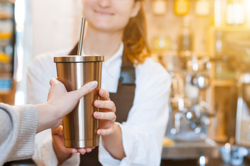 Closeup female hands are holding metal reusable tumbler cup mug of coffee. Woman  prepared, brewed espresso, americano, latte cappuccino in cafe. Barista is giving order to client. Take away, to go.