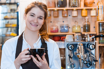 Young woman barista in brown apron is drinking coffee on workplace. Girl is waiting for client in cafe, restaurant. Professional machine for preparing brewing americano, espresso on background.