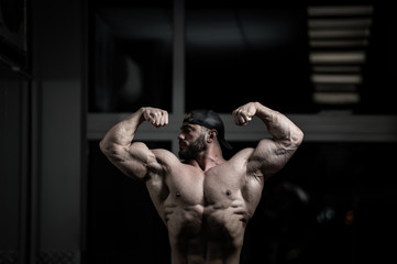 handsome active strong bearded caucasian man in cap showing double biceps muscles in dark fitness gym during training workout