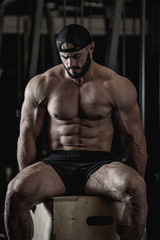 Fototapeta na wymiar strong handsome young muscular male with beard and powerful abdominal muscles wearing cap and trunks sitting on wooden box during workout training