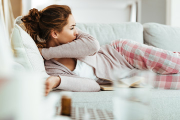Young woman coughing into elbow while lying down on sofa in the living room.