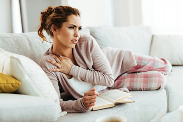 Young woman feeling pain while lying on the sofa and reading a book.