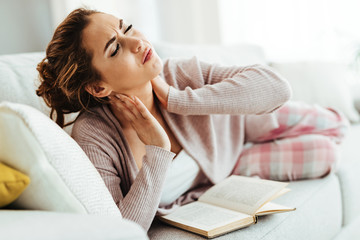 Naklejka premium Young woman having neckache while relaxing on the sofa at home.
