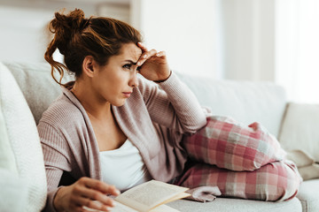 Young woman having headache while reading book on the sofa at home.