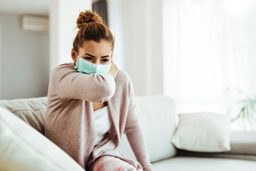 Sick woman with face mask sneezing into elbow at home.