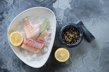 White bowl with fresh codfish fillet on ice and seasonings, top view on a grey concrete background,...