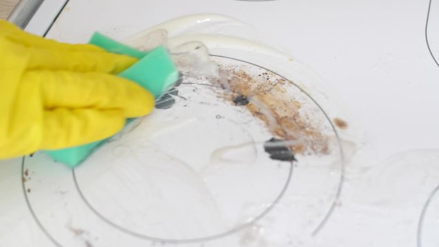 Hand in yellow rubber glove washes dirty induction stove with a sponge in kitchen. Housekeeping concept. Quarantine work at home. Stay home. Hands washes a stove with a cleaner. House cleaning. 