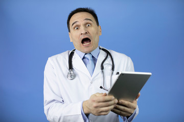 Portrait of a scared adult male physician with stethoscope dressed in white medical coat holding a tablet. Isolated blue background. The man opened his mouth wide and wonders.